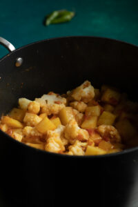 A black pot filled with Pakistani Aloo Gobi-cooked cauliflower and potato pieces in a spiced tomato sauce, with a blurred green chili in the background on a teal surface.