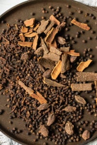 A brown plate filled with whole spices-black peppercorns, dried cloves, cinnamon sticks, and large black cardamom pods-perfect for creating authentic Pakistani Garam Masala, all arranged in a loose pile.