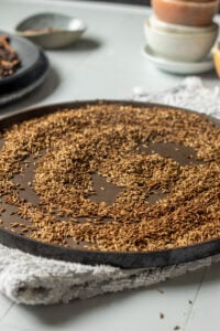 A round black tray filled with dried cumin seeds, a key ingredient in Pakistani Garam Masala, spread evenly on a textured kitchen towel atop a light gray countertop, with bowls and trays blurred in the background.