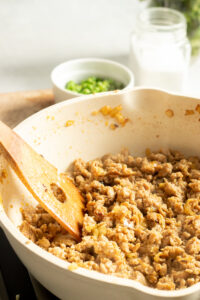 A skillet filled with browned Chicken Keema and sautéed onions is being stirred with a wooden spatula. In the background, theres a small bowl of chopped green onions and a jar of milk.