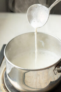 A ladle pours hot, steaming milk for Doodh Patti into a stainless steel pot on a stovetop. The milk is frothy, with wisps of steam rising above the pot.