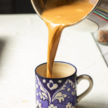 A metal pot pours creamy Doodh Patti chai into a blue and white patterned mug on a white textured surface. Some tea splashes on the table.