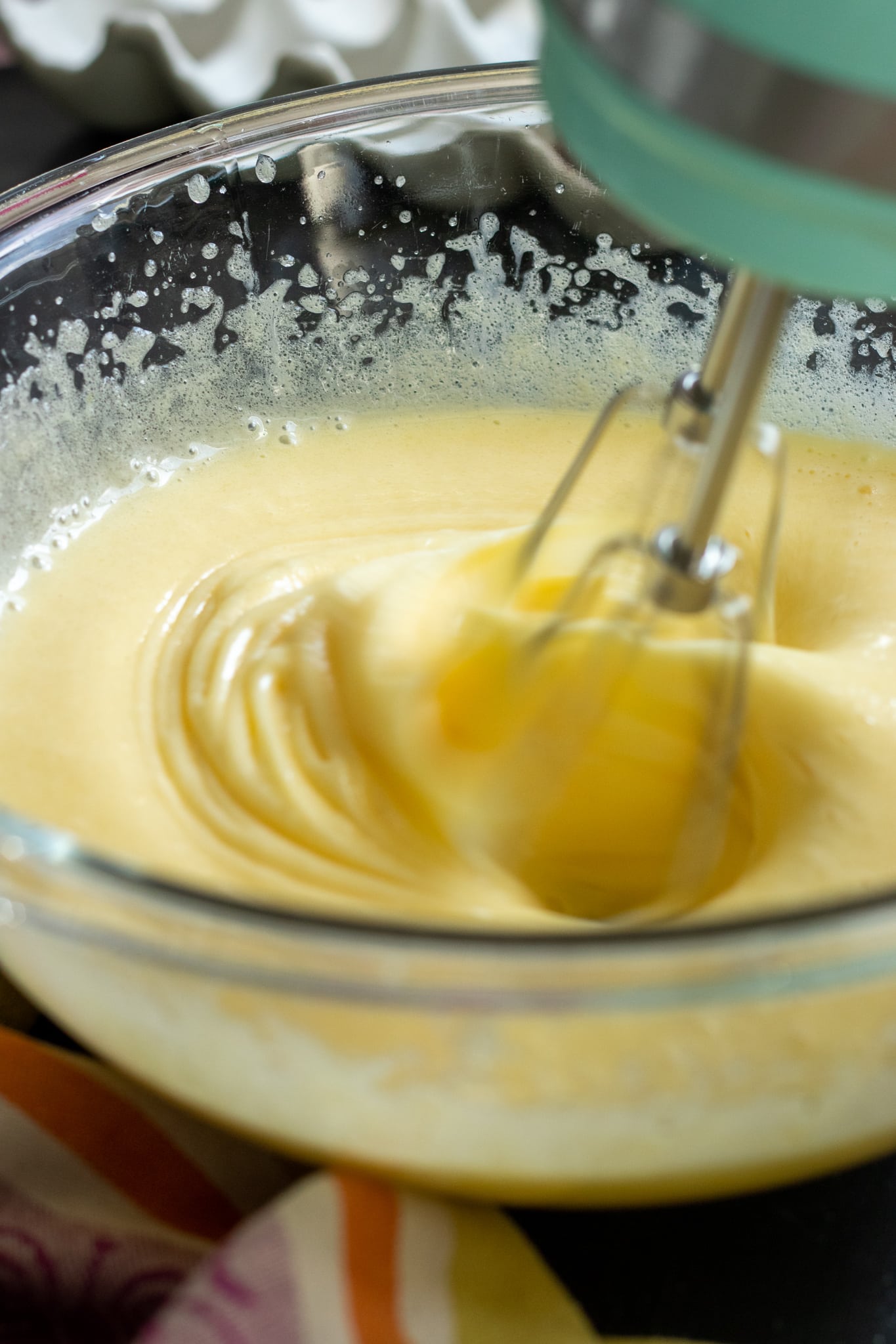 A hand mixer blends a creamy yellow Marble Cake with Oil batter in a clear glass bowl, creating smooth, swirling patterns.