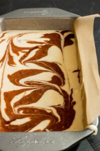 A rectangular metal baking pan lined with parchment paper contains a marbled mixture of chocolate and vanilla Marble Cake with Oil, swirled together in a decorative pattern.
