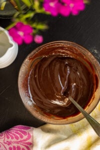 A bowl of smooth, glossy chocolate ganache with a spoon in it, placed on a dark surface near a pink and yellow patterned cloth and blurred pink flowers-perfect for topping your favorite marble cake with oil.
