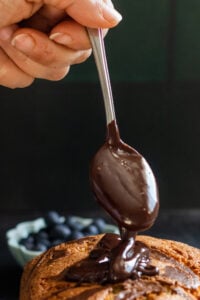 A hand holds a spoon dripping melted chocolate onto a stack of pancakes, reminiscent of marble cake with oil, with a bowl of blueberries blurred in the background.