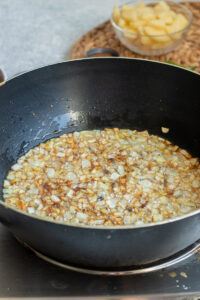 Chopped onions are being sautéed and browned in oil in a black frying pan on a stovetop, the perfect start for Aloo Keema. In the background, a bowl of diced potatoes sits on a woven mat.