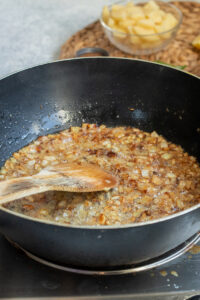 Chopped onions are being sautéed in a black frying pan with oil and stirred with a wooden spoon—a key step in preparing Aloo Keema. In the background, there is a glass bowl with chopped potatoes.