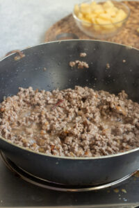 Ground beef is being cooked in a black skillet on a stovetop for a classic Aloo Keema, with steam rising from the qeema. In the background, a bowl of sliced potatoes sits slightly out of focus.