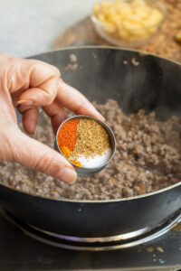 A hand holds a small bowl with three types of ground spices over a pan of browning Qeema for Aloo Keema on a stovetop. In the background, a bowl of sliced ingredients is partially visible.