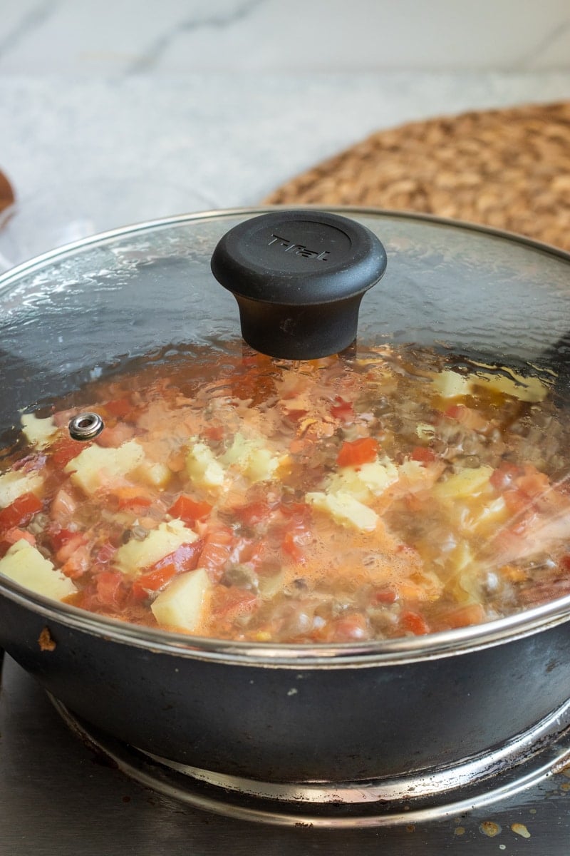 A pot with a glass lid simmers a savory mixture like Aloo Keema, diced tomatoes, vegetables, and chunks of butter or cheese on the stovetop. Steam and condensation gather on the lid as it cooks.