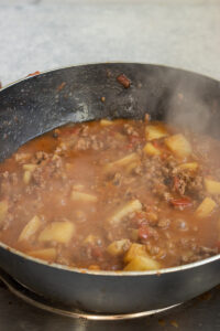 A steaming pan filled with a hearty Aloo Keema stew, featuring ground meat, potatoes, tomatoes, and beans, cooking on a stovetop.