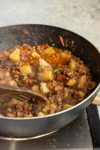 Ground beef and diced potatoes simmer in a tomato-based sauce inside a deep skillet, creating a classic Aloo Keema. A wooden spoon stirs the mixture as steam rises from this hot, hearty qeema dish.