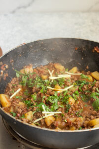 A pan filled with spiced ground meat and potato chunks—classic Aloo Keema—in a rich tomato-based sauce, garnished with fresh chopped cilantro and thin slices of ginger. Steam rises from this hot qeema dish.