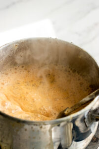 A stainless steel pot filled with bubbling, frothy Doodh Patti is boiling on a stove. Steam rises from the surface, and a metal spoon rests inside the pot. The background shows a white countertop.