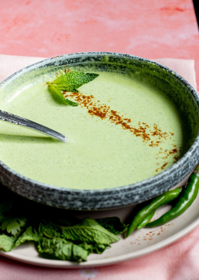 A bowl of creamy green soup, reminiscent of Pudina Raita, is garnished with a mint leaf and a sprinkle of spice. Green chilies and fresh herbs are on the side, all set on a pink surface with a spoon resting inside.