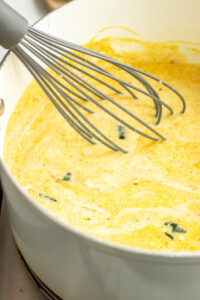 A metal whisk stirs a creamy yellow Kadhi Pakora soup with green herb pieces in a white pot.