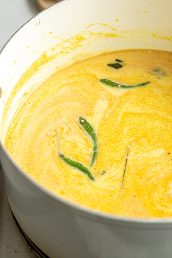 A white pot filled with creamy yellow karhi, with green chili peppers and seasonings visible on the surface.