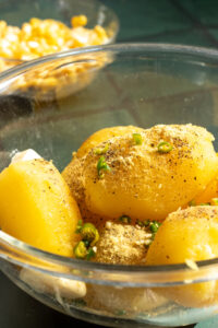 A glass bowl filled with whole, peeled boiled potatoes seasoned with ground spices, chopped green chilies, and black pepper-perfect for preparing Aloo Cutlets or following your favorite Potato Cutlets Recipe. A blurred bowl of corn kernels sits in the background.