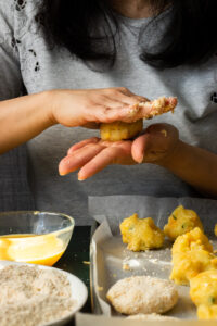 A person shapes dough with their hands over a tray lined with parchment paper. Nearby are a bowl of beaten eggs and breadcrumbs, hinting at the classic preparation of Aloo Cutlets or following a Potato Cutlets Recipe.
