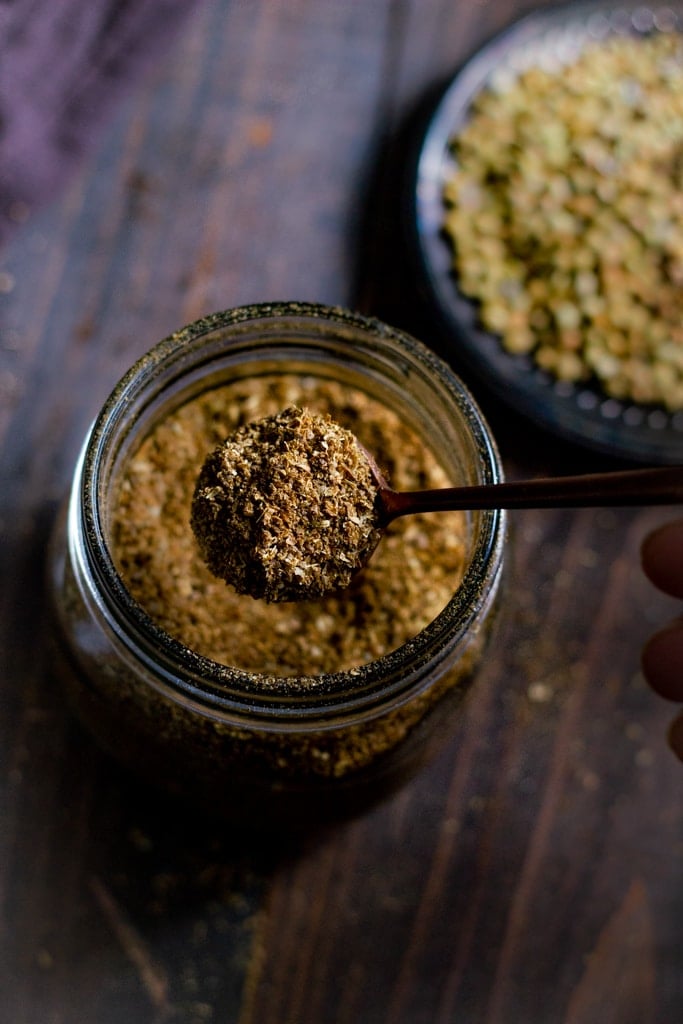 top view of a jar of ground roasted coriander powder with a spoon in it. a dish of coriander seeds in the background