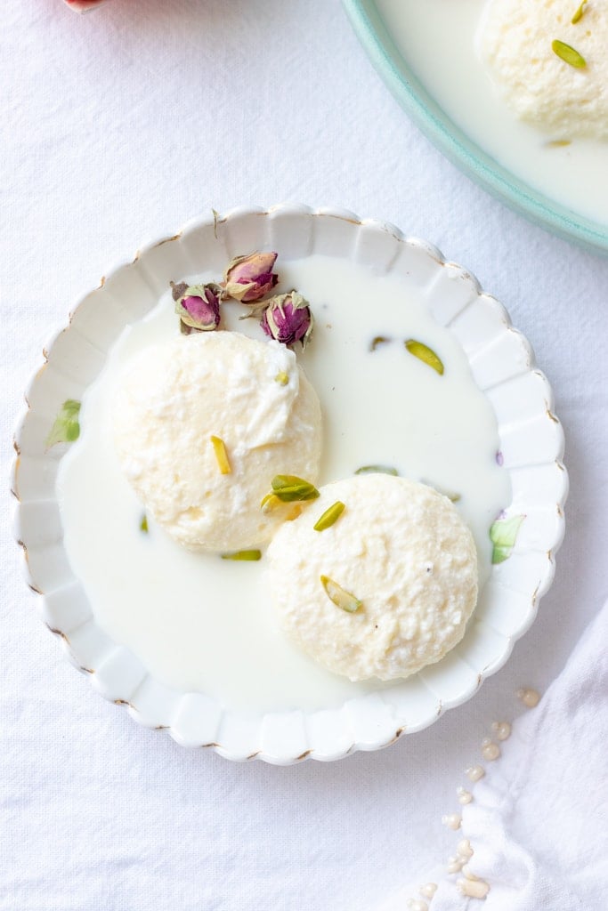 2 ras malai pieces in a plate with milk, a dried rose and pistachios for garnish