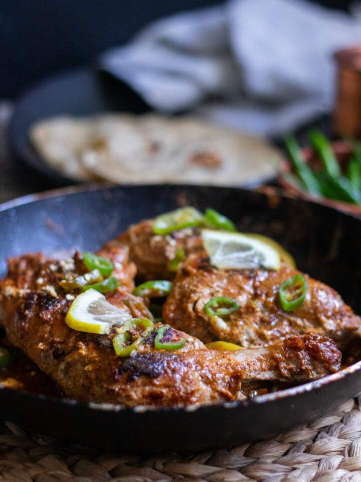 two cooked pieces of tawa chicken in a pan, roti in the backrgound