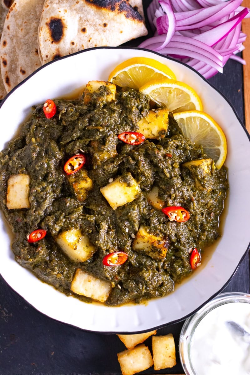 top down view of palak paneer on a board with plain yoghurt, roti, and paneer cubes