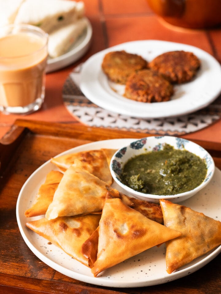a plate of samosas and chutney on a tray with shami kabab and chai in the background