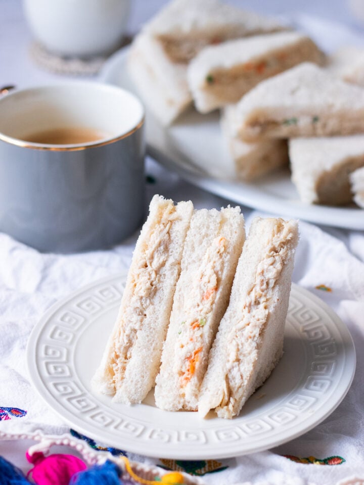creamy bakery style mayo chicken sandwiches on a plate, cup of tea and a platter of sandwiches in the background