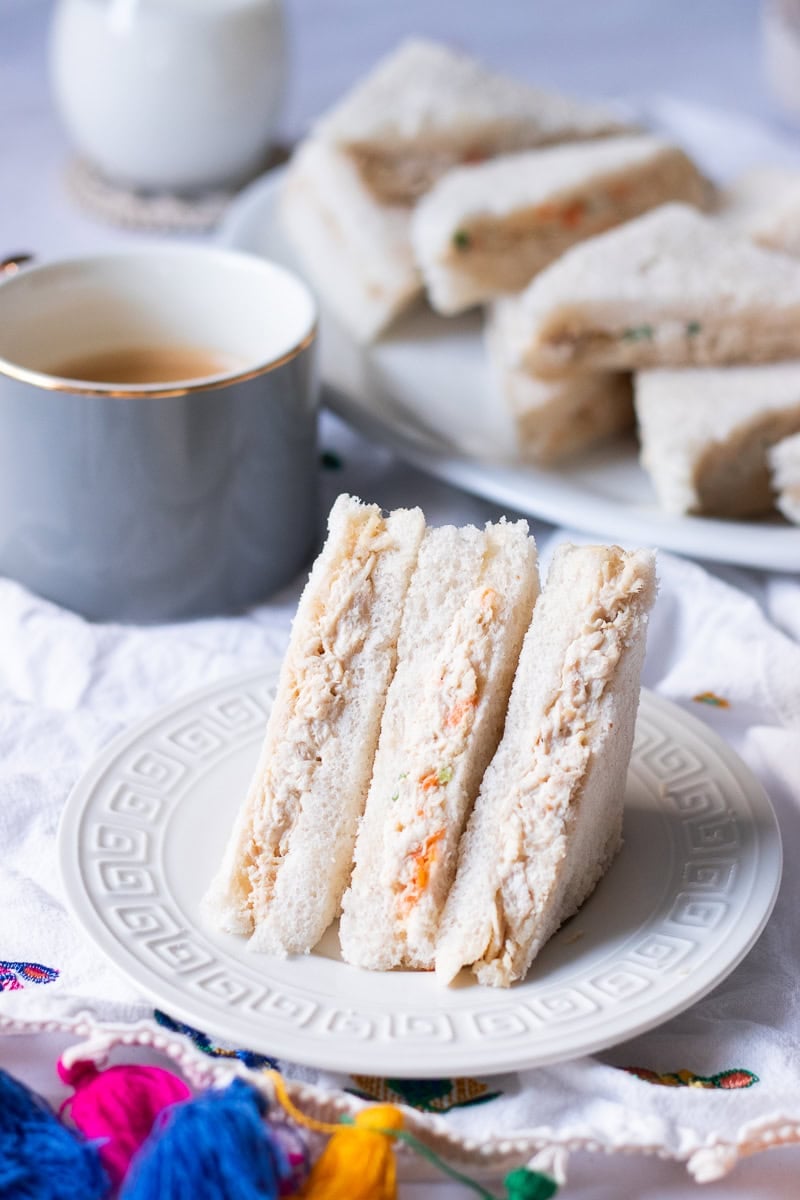 creamy bakery style mayo chicken sandwiches on a plate, cup of tea and a platter of sandwiches in the background
